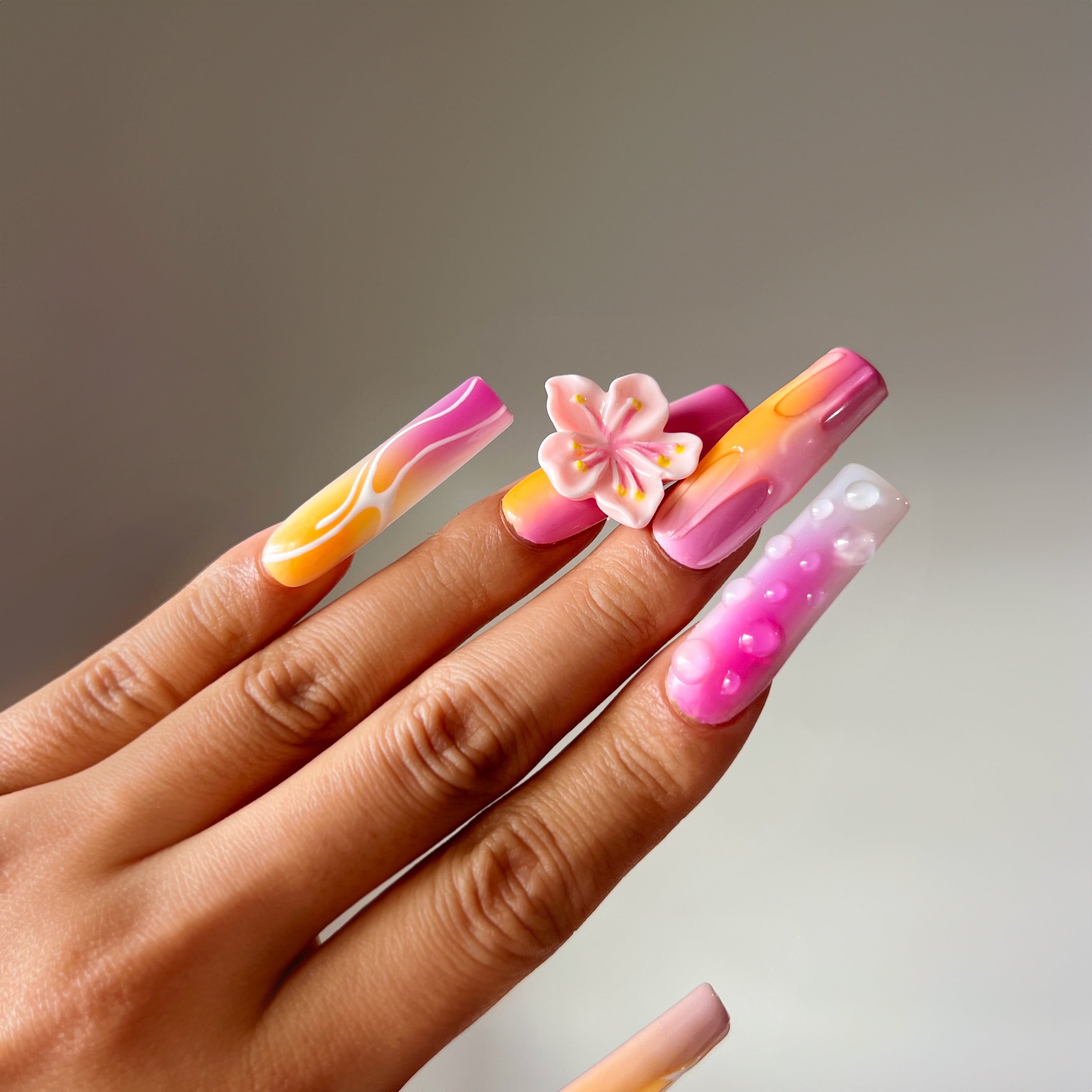 Extra-long square press-on nails with pink and orange gradient, 3D flowers, white accents, and water drop details for a vibrant, floral design.