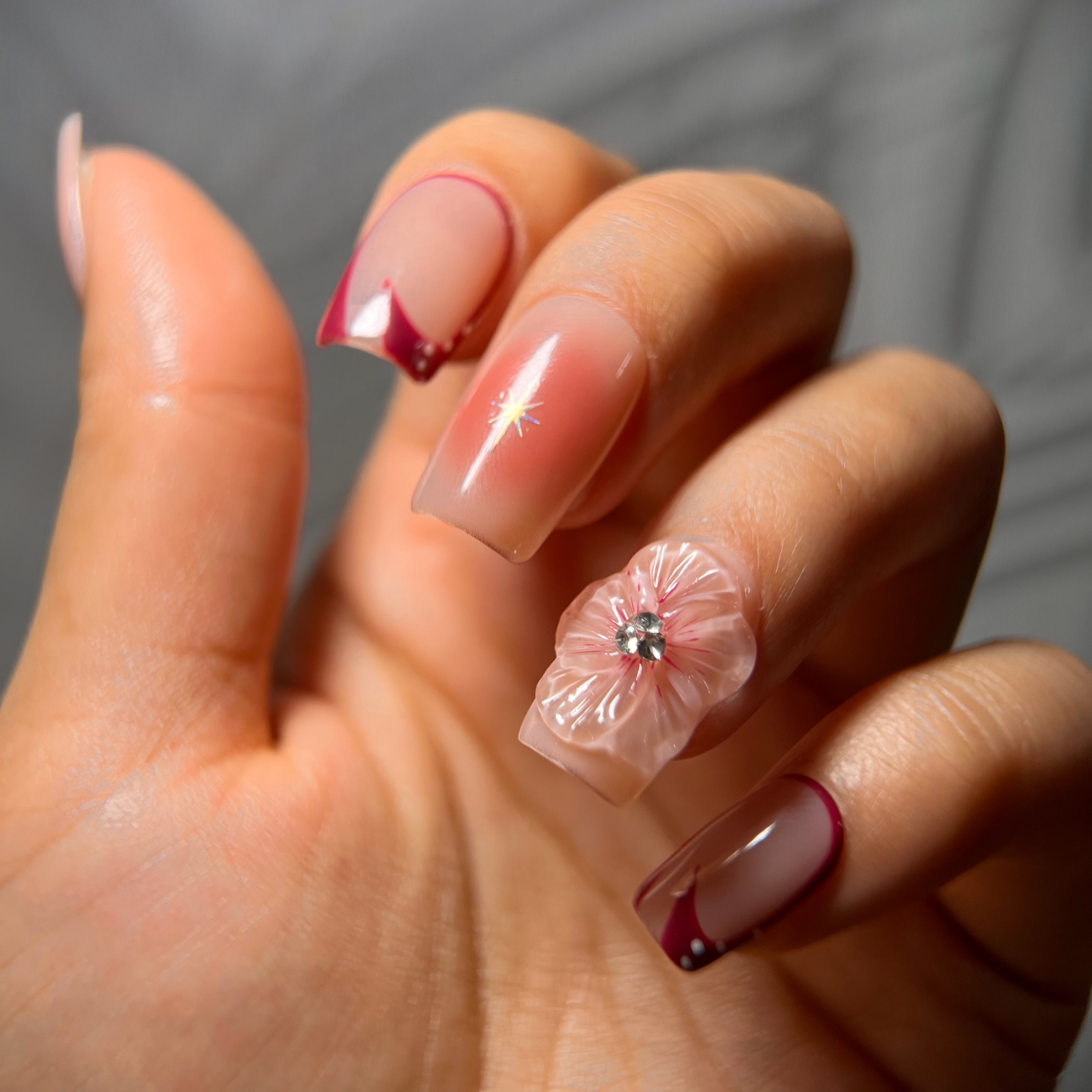 Nude press-on nails with burgundy airbrush, hibiscus flowers, and metallic charms for a bold floral design.